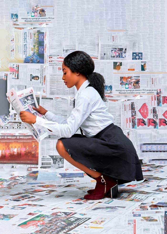 Young woman reading newspapers surrounded by walls and floor covered in newspapers.
